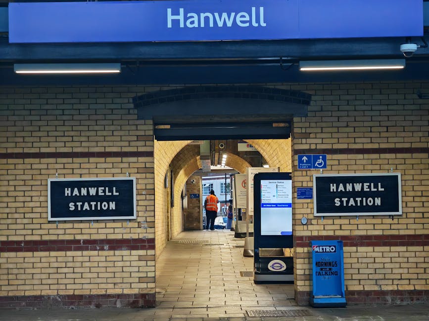 The entrance to Hanwell Station is framed by yellow brick walls and archways, with a blue sign displaying 'Hanwell' mounted above the arch. Two black-and-white signs reading 'Hanwell Station' are posi